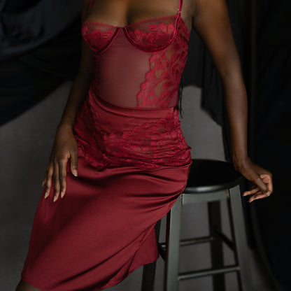 Woman wearing a red lace dress layered over a red lace bodysuit sitting on a stool against a dark background