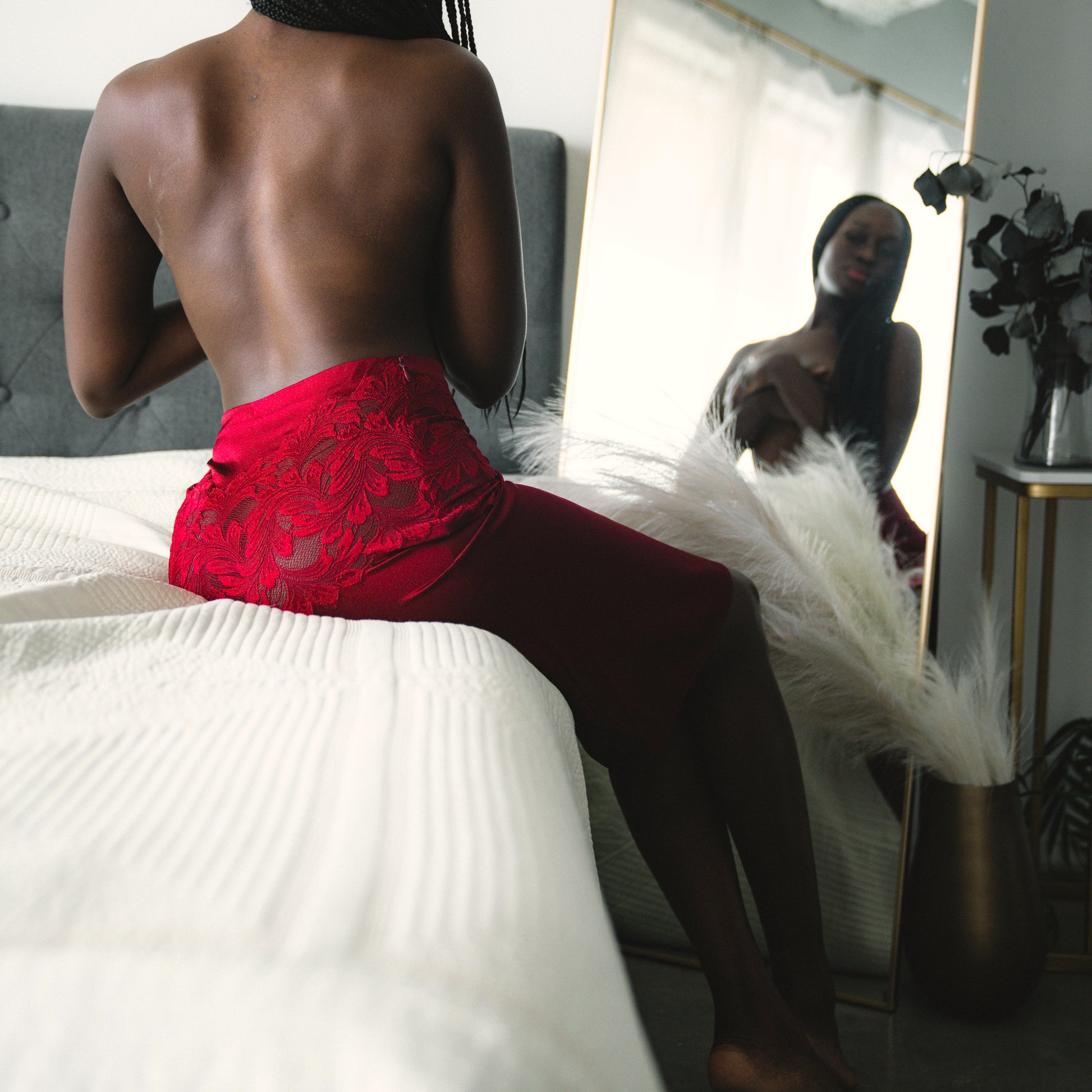 Woman wearing red lace skirt sitting on a bed in front of a mirror.